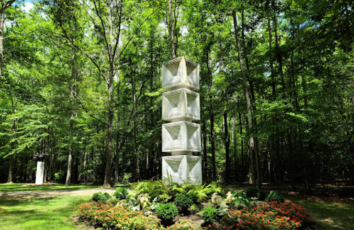 A tall, white sculpture with geometric shapes stands amidst lush green trees and colorful flower beds.