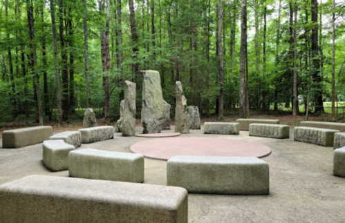 A circular stone gathering area surrounded by trees, featuring large rock formations and curved stone benches.