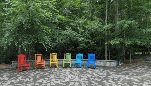 A row of colorful rocking chairs in red, orange, yellow, green, blue, and purple, set against a lush green forest backdrop.