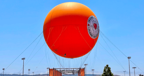 A large orange hot air balloon with a heart logo floats against a clear blue sky.