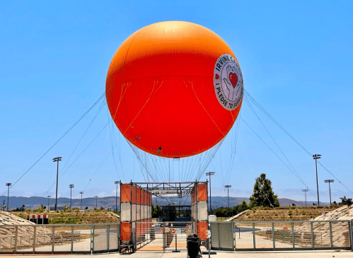 A large orange hot air balloon with a logo floats above a fenced area on a clear day.