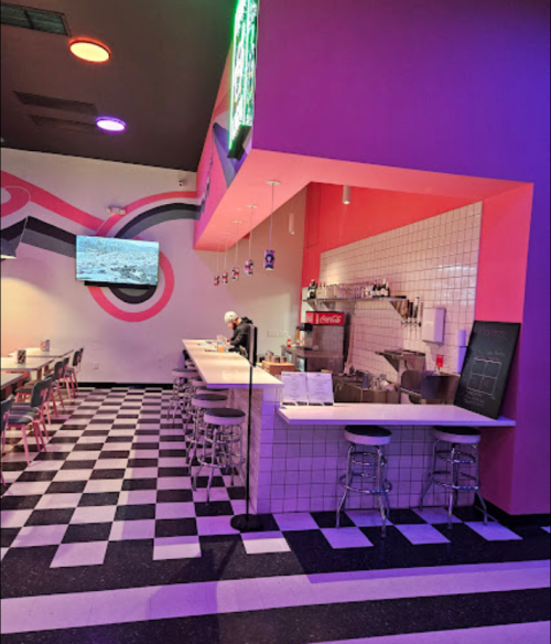 A colorful diner interior with a checkered floor, bar seating, and a small kitchen area. Neon lights add ambiance.