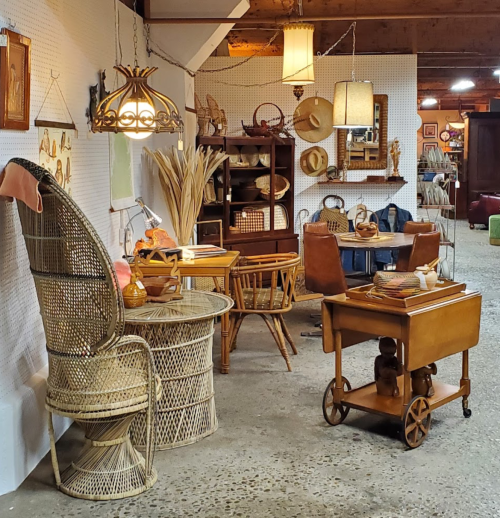 A vintage-inspired room featuring wicker furniture, a wooden cart, and various decorative items on display.