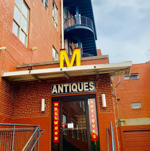 A brick building with a large yellow "M" sign and "Mellwood Antiques" displayed above the entrance.