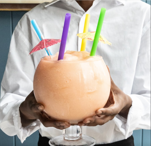 A person holds a large, fruity drink with colorful straws and tiny umbrellas, set against a blue background.