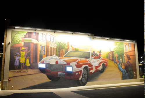 A vibrant mural at night depicting a classic car with characters, set against a colorful urban backdrop.