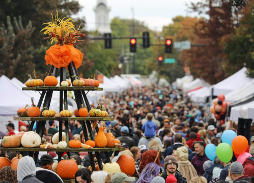 A bustling street festival with a large pumpkin display in the foreground and crowds of people enjoying the event.