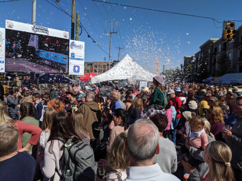 A lively crowd gathers at an outdoor event, with confetti falling and a festive atmosphere under a clear blue sky.