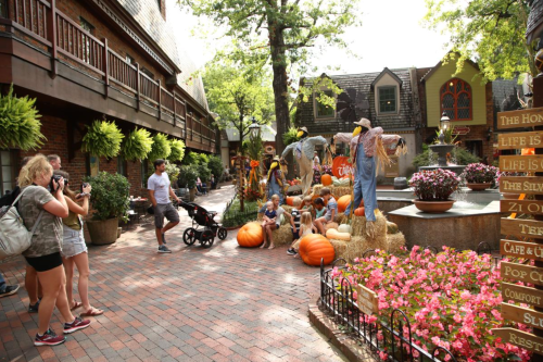 A festive outdoor scene with people taking photos, pumpkins, hay bales, and autumn decorations in a charming plaza.
