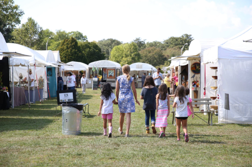 A woman and four children walk through an outdoor market with white tents and various displays.