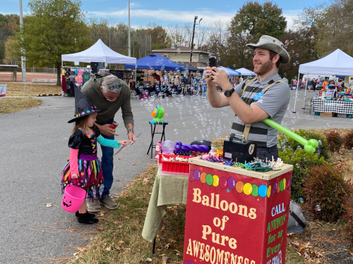 A vendor in a hat blows bubbles while a child in a costume watches, with colorful balloons and a sign nearby.