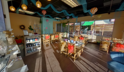 Brightly lit café with wooden tables, colorful decorations, and a cozy atmosphere, featuring plants and hanging lanterns.