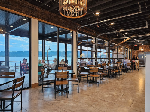 A spacious restaurant with large windows overlooking a lake, featuring wooden decor and tables with diners.