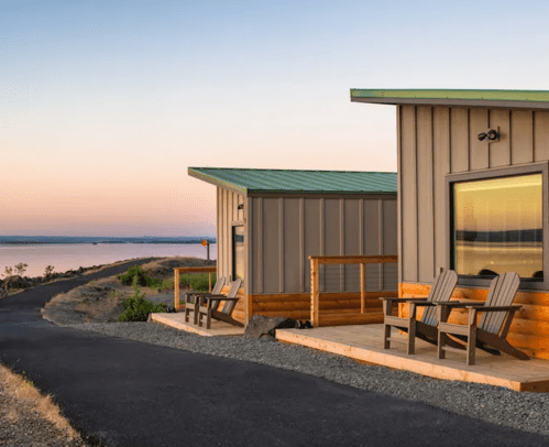 Two modern cabins with wooden decks overlook a serene landscape at sunset, with a path leading to them.
