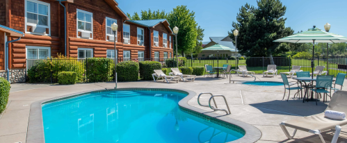 A sunny pool area with lounge chairs, umbrellas, and a log cabin-style building in the background.