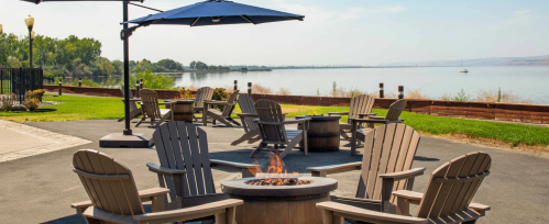 A lakeside patio with wooden chairs, a fire pit, and an umbrella, overlooking calm water and greenery.