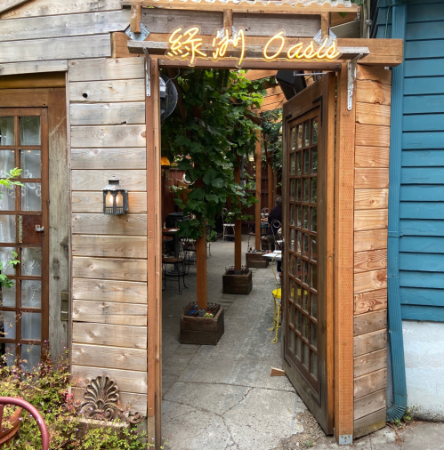 A wooden entrance with a sign reading "Oasis" leads into a cozy outdoor dining area surrounded by greenery.