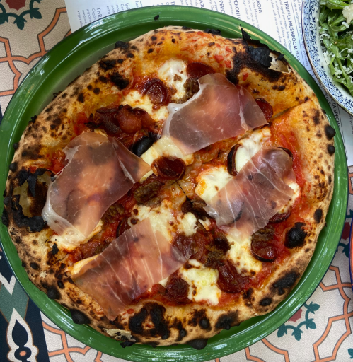 A close-up of a pizza topped with melted cheese, slices of prosciutto, and pepperoni on a charred crust.