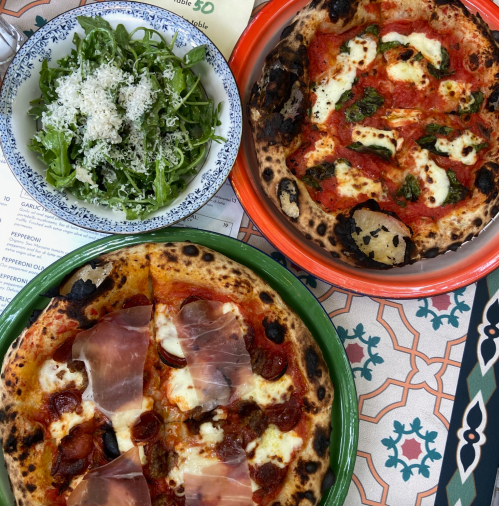 Two pizzas on colorful plates, one topped with pepperoni and prosciutto, the other with mozzarella and basil, alongside a salad.