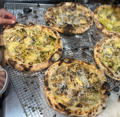 Freshly baked pizzas on a wire rack, topped with various vegetables and cheese, with a hand reaching for one.