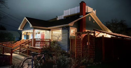 A cozy blue house at night, illuminated with warm lights, featuring a wooden deck and a bicycle parked outside.