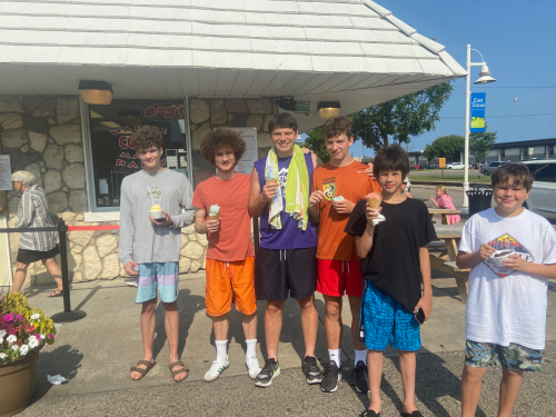 A group of six boys stands outside an ice cream shop, each holding ice cream cones and smiling.