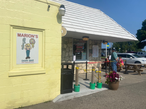 A yellow building with a cartoon sign for "Marion's Dairy Bar" and a man waiting outside near picnic tables.