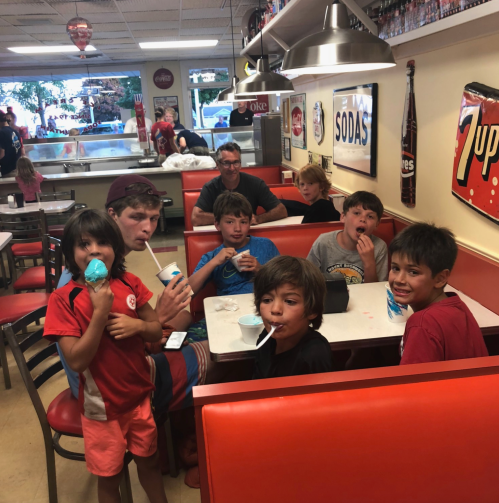 A group of children and an adult sit in a diner, enjoying ice cream and snacks at a red booth.