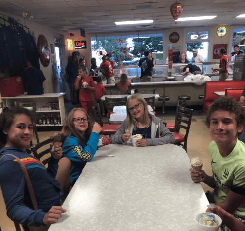 Four children enjoy ice cream at a busy ice cream shop, with others in the background.