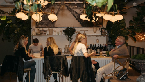 A group of people socializing at a bar surrounded by plants and warm lighting, with bottles of wine in the background.