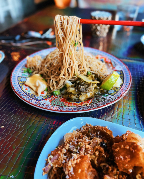 A plate of noodles with vegetables, garnished with lime, and a side of crispy dish, all on a colorful table.
