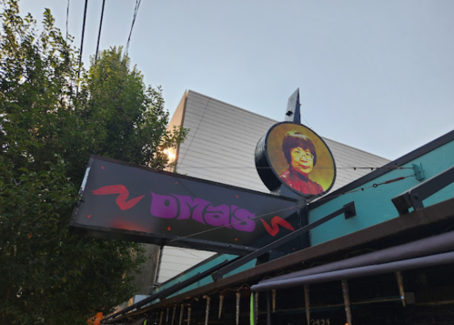 Sign for "Dya's" featuring a portrait of a woman, set against a clear sky and trees in the background.