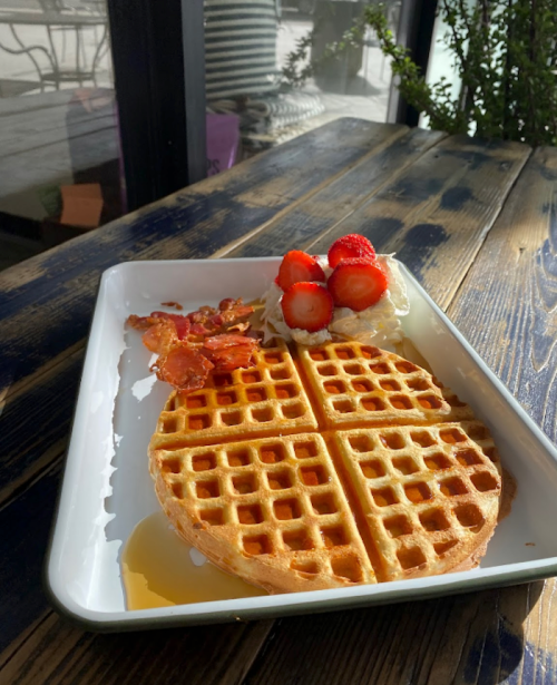 A plate of waffles topped with strawberries, whipped cream, and bacon, served with syrup on a wooden table.