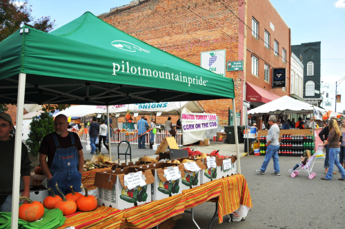 A bustling farmers market with a green tent, pumpkins, and vendors selling fresh produce in a small town setting.