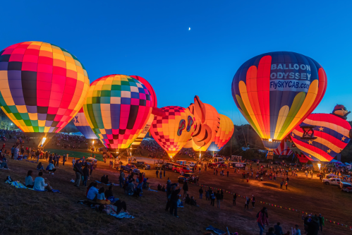 Colorful hot air balloons illuminated at dusk, with a crowd gathered below and a crescent moon in the sky.