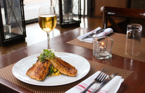 A plate of grilled fish with vegetables, accompanied by a glass of white wine, set on a wooden table in a cozy restaurant.