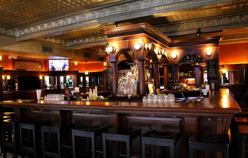 A wooden bar with high stools, shelves of liquor, and warm lighting in a cozy pub setting.