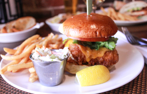A crispy chicken sandwich with lettuce, tomato, and cheese, served with fries and coleslaw in a metal cup.