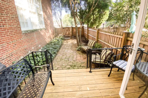 A cozy outdoor patio with wooden decking, black chairs, and a brick pathway surrounded by greenery and a wooden fence.