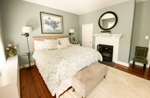 Cozy bedroom with a large bed, decorative pillows, a fireplace, and a mirror, featuring soft blue walls and wooden floors.