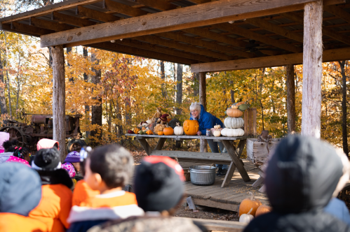 A person demonstrates pumpkin carving to a group of children under a wooden shelter surrounded by autumn foliage.