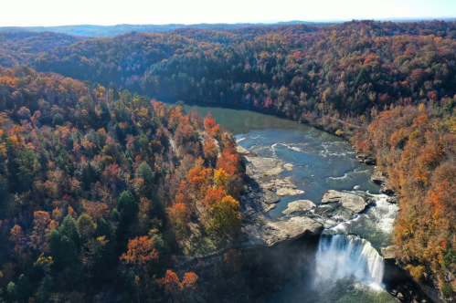 Aerial view of a waterfall surrounded by vibrant autumn foliage and a winding river in a forested landscape.