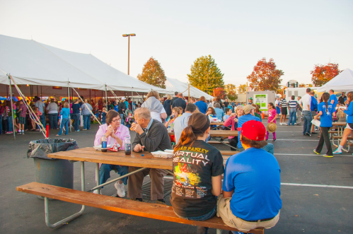 A lively outdoor event with people socializing at picnic tables under tents, surrounded by autumn trees.