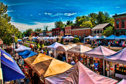 A vibrant outdoor market scene with colorful tents, vendors, and crowds under a blue sky with scattered clouds.