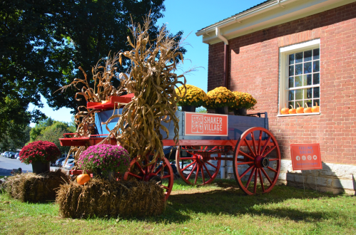 A decorated wagon with pumpkins and flowers, surrounded by hay bales, outside a brick building in a village setting.