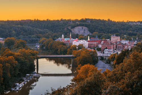 A scenic view of a river town at sunset, with autumn foliage and historic buildings along the water's edge.
