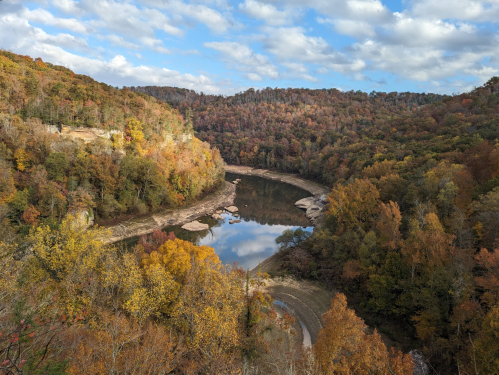 A serene river winding through a forest of autumn-colored trees under a partly cloudy sky.