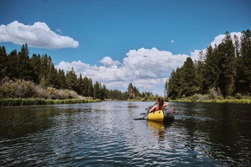 A person in a yellow kayak paddles on a calm river surrounded by lush trees and a blue sky with fluffy clouds.