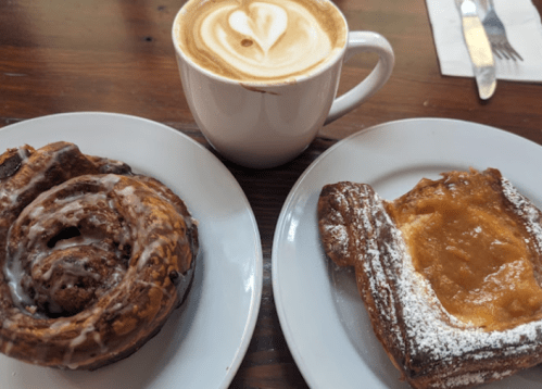 A cinnamon roll and a pastry with fruit filling, accompanied by a latte with a heart-shaped foam design.