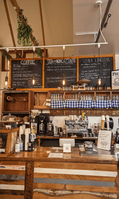 A cozy café counter with a chalkboard menu, wooden shelves, and hanging lights, showcasing coffee and tea options.